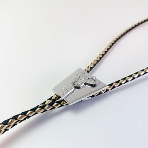May include: A silver bolo tie with a braided black and gold cord. The tie features a square-shaped pendant with an intricate design.