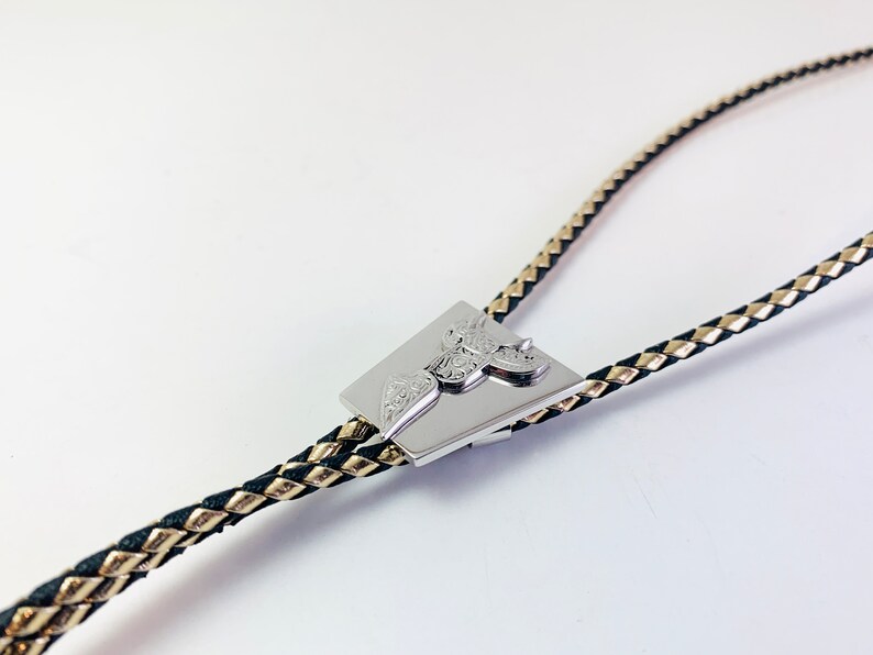 May include: A silver bolo tie with a braided black and gold cord. The tie features a square-shaped pendant with an intricate design.