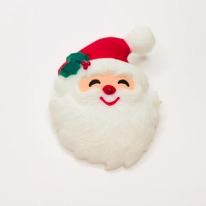 May include: A Christmas decoration in the shape of Santa Claus's face. The face has a red nose and a smiling red mouth. The Santa hat is red with a white bobble and green holly. The beard is white and fluffy.