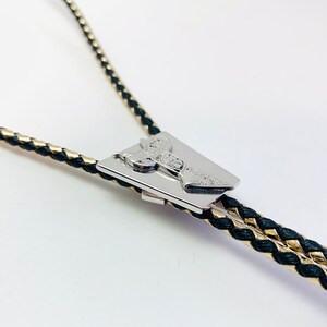 May include: A black and gold braided bolo tie with a silver, rectangular, engraved pendant.