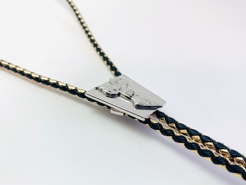 May include: A black and gold braided bolo tie with a silver, rectangular, engraved pendant.