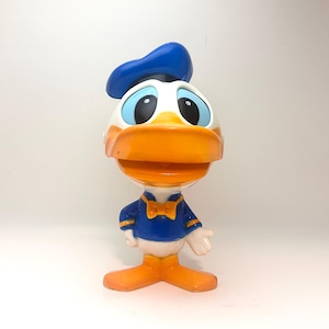 May include: A blue and white plastic toy figurine of Donald Duck wearing a blue sailor hat and a blue bow tie. The figurine has a yellow beak and orange feet.