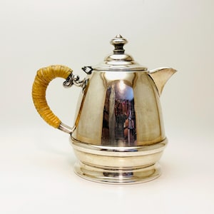 May include: A silver teapot with a woven, yellow handle. The teapot has a rounded body and a lid with a finial.