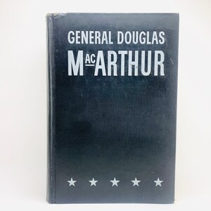以下が含まれることがあります： 黒い表紙に白い文字で「GENERAL DOUGLAS MACARTHUR」と書かれたハードカバーの本。下部には5つの白い星が並んでいます。白い背景に本が置かれています。