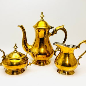 May include: A gold-tone tea set comprising a teapot, creamer, and sugar bowl. The set features a traditional design with ornate handles and a polished finish.