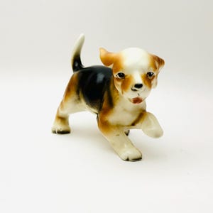 May include: A ceramic figurine of a brown, black and white puppy with its tongue sticking out. The puppy is standing on all fours and has its right paw raised.