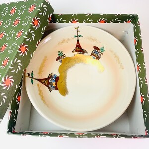 May include: A white ceramic dish with a gold rim and a hand-painted design of three houses on a hill. The dish is in a green and white box with a swirling pattern.