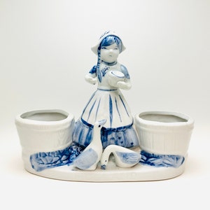 May include: A blue and white ceramic planter featuring a Dutch girl figurine holding a pitcher and two geese. The planter has two separate pots for plants.