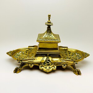 May include: An antique brass inkwell, featuring a central compartment and two side trays. The inkwell has detailed ornamentation, including a small, pagoda-style top and filigree patterns. It is supported by four claw feet.