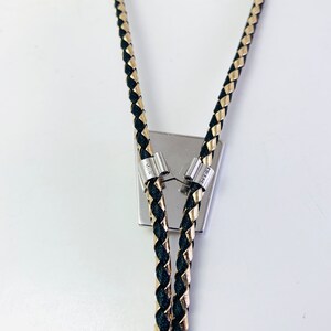 May include: Black and gold braided bolo tie with silver tips. The bolo tie is attached to a silver square pendant.