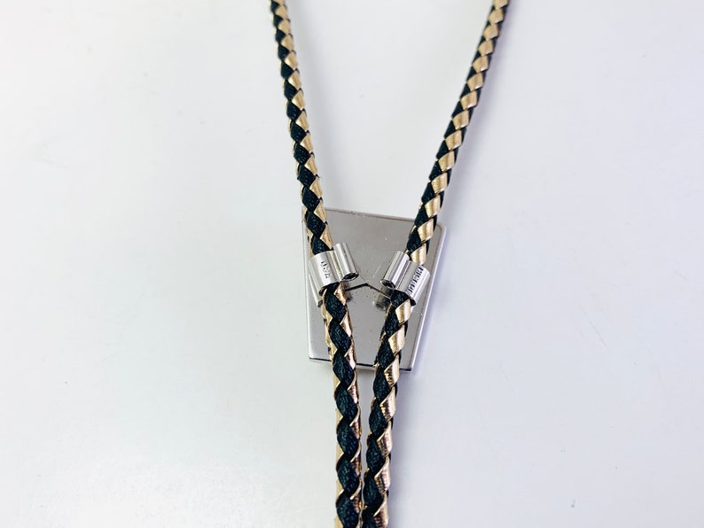 May include: Black and gold braided bolo tie with silver tips. The bolo tie is attached to a silver square pendant.