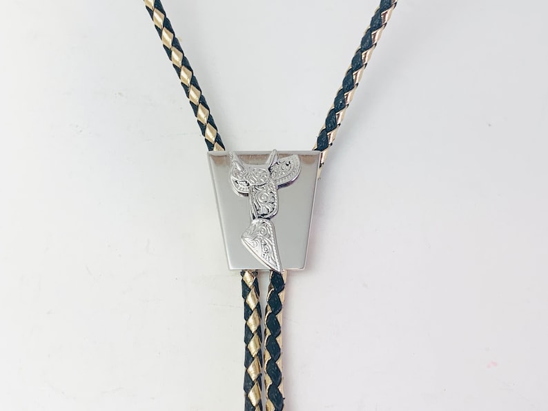 May include: A silver bolo tie with a black and gold braided cord. The tie features a silver charm in the shape of a saddle.