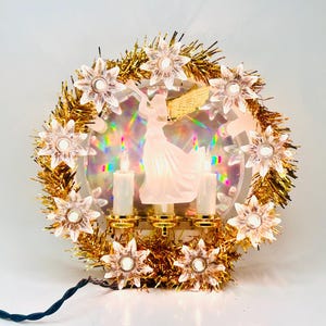 May include: A circular Christmas decoration featuring a gold tinsel wreath adorned with clear, star-shaped light covers. In the centre, a translucent angel figure stands between two white candle-shaped lights against a holographic background.