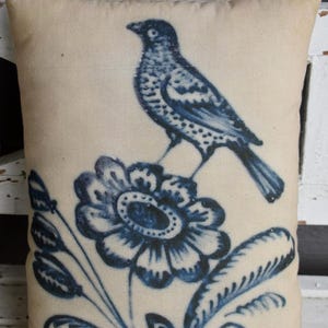 May include: Rectangular beige pillow with a blue bird perched on a blue flower. The bird and floral design are in a vintage, folk art style. The pillow is made of a soft fabric and would be a decorative accent for a bed or sofa.