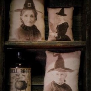 May include: Five vintage-style fabric pillows with images of women wearing witch hats. The pillows are arranged on a wooden shelf with a bottle of liquid and a lit candle.