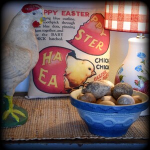 May include: A decorative Easter-themed display featuring a ceramic chicken figurine, a pillow with a chick graphic, and a blue bowl filled with stones. A lamp with a red and white checkered shade is also visible.