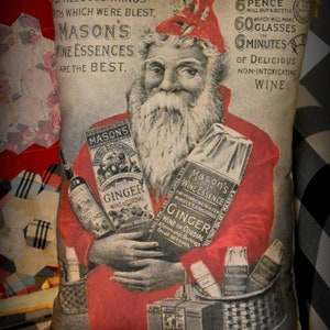 May include: A vintage pillow featuring a Santa Claus illustration holding bottles of Mason's Wine Essences. The pillow is made of a beige fabric with red and black checkered trim. The text on the pillow reads "Of all good things with which we're blest, Mason's Wine Essences are the best." and "6 pence will buy a bottle which will make 60 glasses in 6 minutes of delicious non-intoxicating wine."
