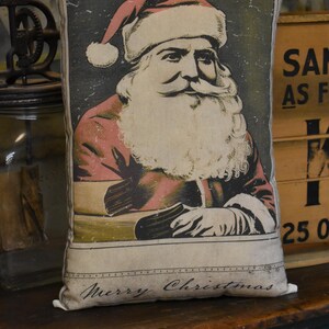 May include: A beige pillow with a vintage illustration of Santa Claus on the front. The illustration is in shades of red, white, and black. The pillow has the text "Merry Christmas" printed on the bottom.