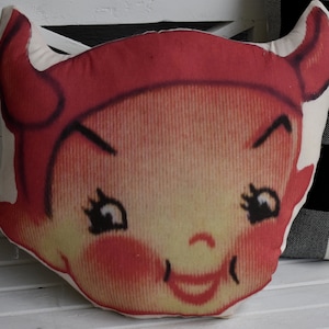 May include: Red and white plush pillow with a smiling devil face. The devil has horns, a red hat, and a mischievous expression.