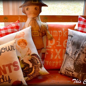May include: A wooden bench with a variety of decorative pillows. The pillows feature Thanksgiving-themed designs, including turkeys, pilgrims, and the text "Get your fat pants ready" and "Pumpkin pie fixin' everyt".