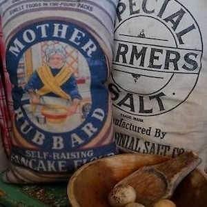 May include: A vintage-style pillow with a blue and white design featuring a woman baking with the text "Mother Hubbard Self-Raising Pancake Flour". The pillow is next to a white sack with the text "Colonial Special Farmers Salt" printed on it.