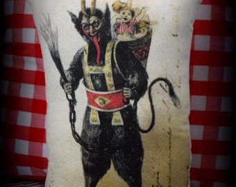 May include: A vintage-style pillow featuring a black and white illustration of a devil holding a whip and carrying a sack of toys. The pillow has a cream-colored background and red and white gingham fabric behind it.