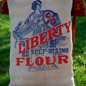 May include: A white pillow with blue and red lettering that reads "LIBERTY SELF-RISING FLOUR". The pillow features a vintage illustration of a woman holding a shield with the letter "S" and the word "MILLING" on it.