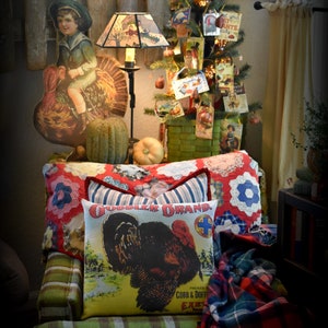May include: A green plaid armchair with a red and white checkered cushion. The armchair has two pillows, one with a red, white, and blue patchwork design and the other with a vintage turkey illustration and the text "Gobbler Brand Packed by Cobb & Doerfler Excelsior, NY". A red and black plaid throw blanket is draped over the arm of the chair. A small Christmas tree decorated with vintage cards and ornaments is in the background.