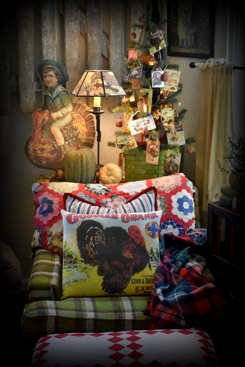 May include: A green plaid armchair with a red and white checkered cushion. The armchair has two pillows, one with a red, white, and blue patchwork design and the other with a vintage turkey illustration and the text "Gobbler Brand Packed by Cobb & Doerfler Excelsior, NY". A red and black plaid throw blanket is draped over the arm of the chair. A small Christmas tree decorated with vintage cards and ornaments is in the background.