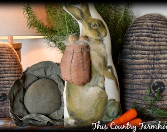 Puede incluir: Una figurita de conejo de estilo vintage con una cesta de huevos, rodeada de una planta verde, una colmena de mimbre marrón y un sombrero de tela gris. El conejo está de pie sobre una mesa con un mantel a cuadros. El texto "This Country Farmhouse" es visible en la parte inferior de la imagen.