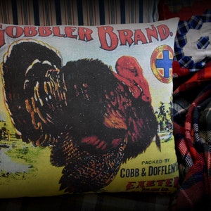 May include: A vintage-style pillow featuring a colorful illustration of a turkey with the text "GOBBLER BRAND. PACKED BY COBB & DOFFLEMYER EXETER, N.H."