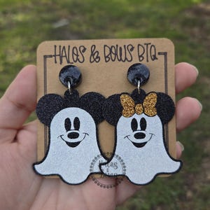 Op de afbeelding: Een paar Halloween-oorbellen met Mickey en Minnie Mouse als geesten. De oorbellen hebben zwarte glitter muizenoren en een witte geestenlichaam. Minnie heeft een gouden glitterstrik. De oorbellen zitten op een bruine kaart met de tekst "HALOS & BOWS BTQ".