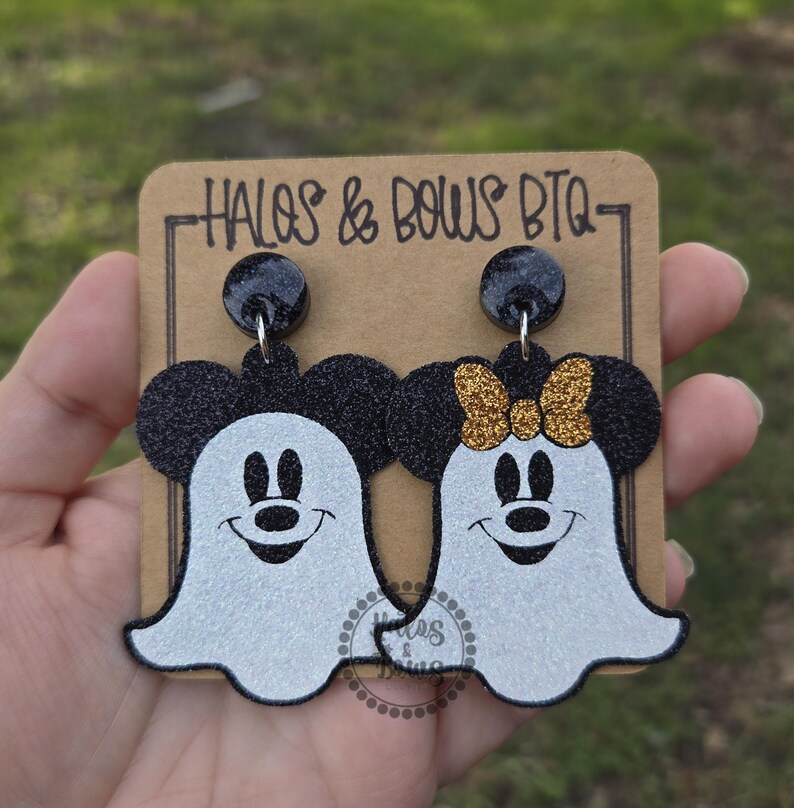 Op de afbeelding: Een paar Halloween-oorbellen met Mickey en Minnie Mouse als geesten. De oorbellen hebben zwarte glitter muizenoren en een witte geestenlichaam. Minnie heeft een gouden glitterstrik. De oorbellen zitten op een bruine kaart met de tekst "HALOS & BOWS BTQ".