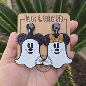 Op de afbeelding: Een paar Halloween-oorbellen met Mickey en Minnie Mouse als geesten. De oorbellen zijn zwart en wit met glitteraccenten en een gouden strik. Ze worden gepresenteerd op een bruine kaart met de tekst "HALOS & BOWS BTQ".
