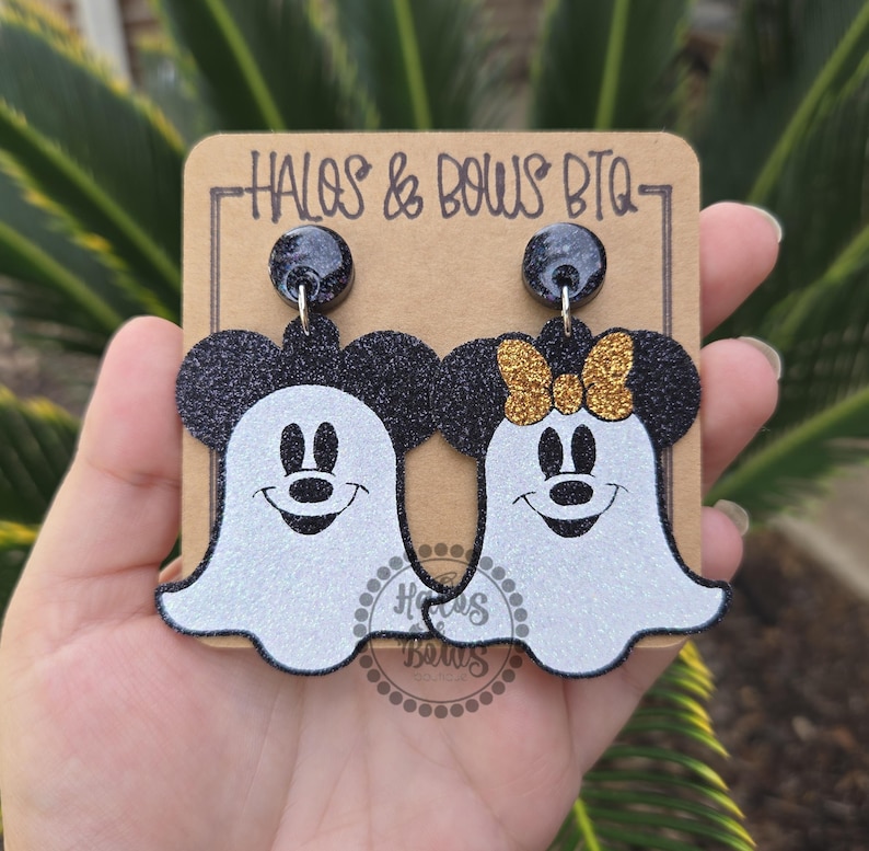 Op de afbeelding: Een paar Halloween-oorbellen met Mickey en Minnie Mouse als geesten. De oorbellen zijn zwart en wit met glitteraccenten en een gouden strik. Ze worden gepresenteerd op een bruine kaart met de tekst "HALOS & BOWS BTQ".