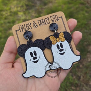 Op de afbeelding: Een paar Halloween-oorbellen met Mickey en Minnie Mouse als geesten. De oorbellen hebben zwarte glitter muizenoren en een witte geestenlichaam met een lachend gezicht. E&eacute;n oorbel heeft een gouden glitterstrik. De oorbellen zitten op een bruine kaart.