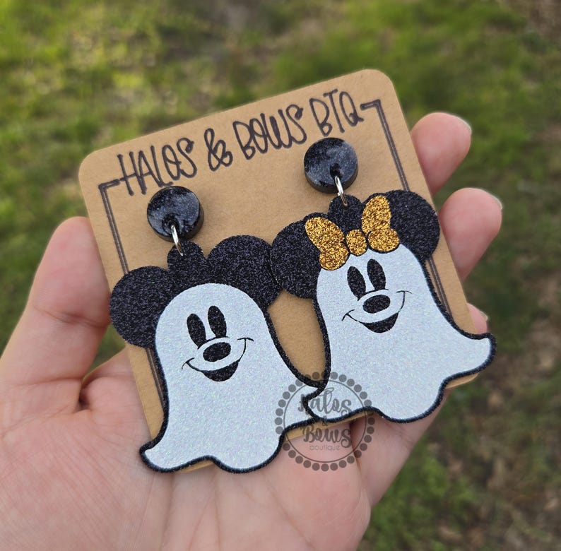 Op de afbeelding: Een paar Halloween-oorbellen met Mickey en Minnie Mouse als geesten. De oorbellen hebben zwarte glitter muizenoren en een witte geestenlichaam met een lachend gezicht. E&eacute;n oorbel heeft een gouden glitterstrik. De oorbellen zitten op een bruine kaart.