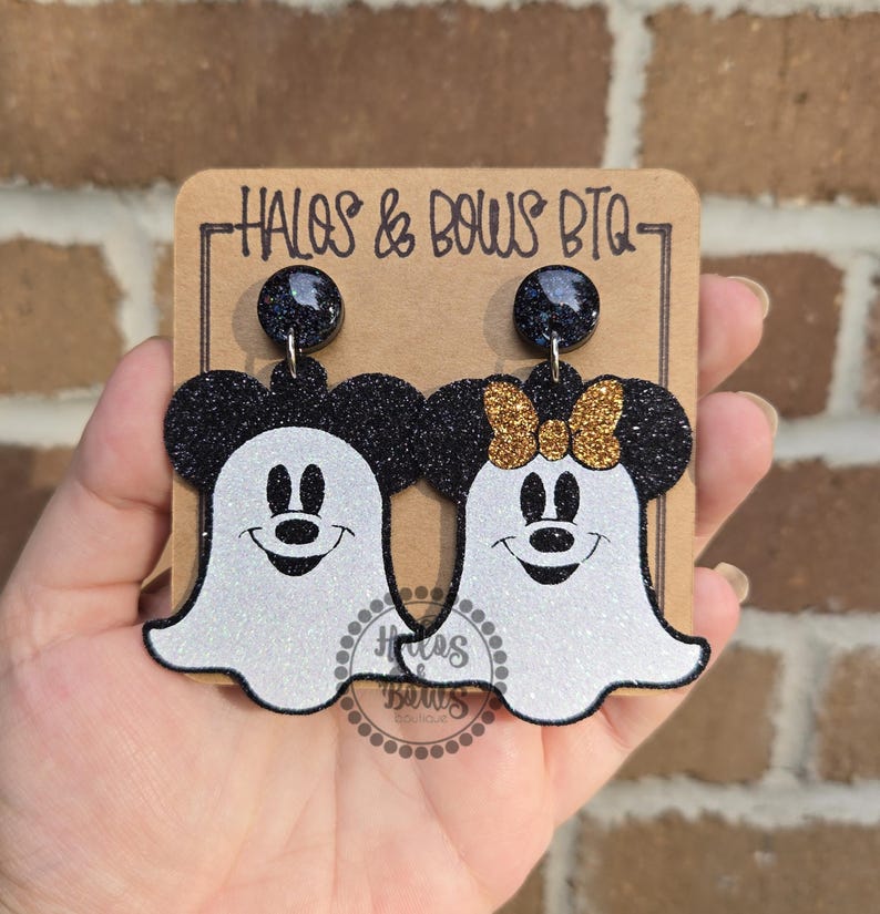 Op de afbeelding: Een paar Halloween-oorbellen met een Mickey Mouse en Minnie Mouse spookontwerp. De oorbellen hebben zwarte glitteraccenten en een witte glitter spooklichaam. De tekst "HALOS & BOWS BTQ" is zichtbaar op de achterkant.