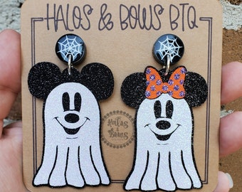 May include: A pair of Halloween-themed earrings featuring Mickey Mouse and Minnie Mouse as ghosts. The earrings are made of black glitter cardstock with white glitter accents. The ghosts have black eyes and a black nose. Minnie Mouse's ghost has a red and white polka dot bow. The earrings are attached to black spiderweb charms.