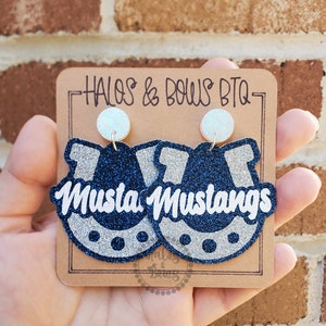 May include: A pair of glittery blue and silver earrings in the shape of a horseshoe with the word "Mustangs" in white. The earrings are held on a brown card with the text "Halos & Bows BTQ".