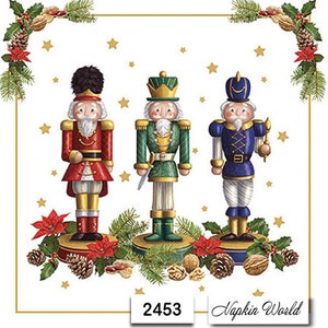 May include: Three nutcracker figurines stand on a white background with a gold border. The nutcrackers are dressed in red, green, and blue uniforms with gold accents. The nutcrackers are surrounded by greenery and poinsettias. The image includes the text "2453" and "Napkin World".