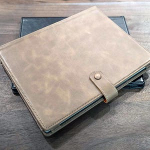 May include: A brown leather portfolio with a black leather flap and a snap closure. The portfolio has a strap with a button closure.