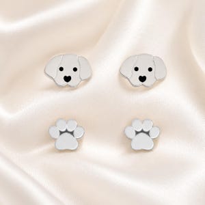 Può includere: Un set di quattro orecchini a lobo color argento. Due orecchini hanno la forma di teste di cane con occhi neri e un naso nero a forma di cuore. Gli altri due orecchini hanno la forma di impronte di zampe con quattro dita arrotondate.