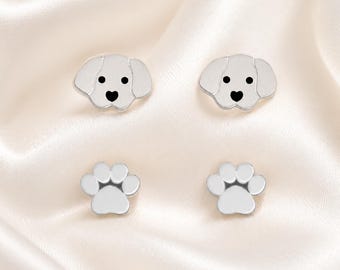 May include: A set of four silver-toned stud earrings. Two earrings are shaped like dog heads with black eyes and a black heart-shaped nose. The other two earrings are shaped like animal paw prints with four rounded toes and a central pad.