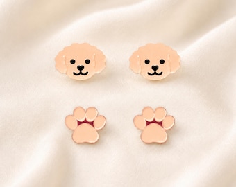 May include: A set of four rose gold-toned stud earrings. Two earrings are shaped like the face of a curly-haired dog with black eyes and a heart-shaped nose. The other two earrings are shaped like a paw print with four distinct rounded toes.