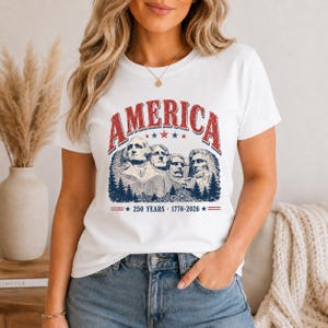 May include: White t-shirt featuring a distressed graphic of Mount Rushmore with the word "AMERICA" in red arched text above. Below the image is the text "250 YEARS - 1776-2026" in red.