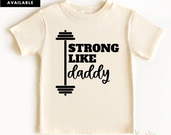 May include: A cream colored t-shirt with a black graphic of a barbell and the text "Strong Like Daddy".
