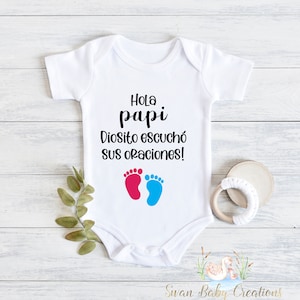 May include: White baby onesie with the text "Hola papi Diosito escuchó sus oraciones!" in black. Below the text are two footprints, one pink and one blue. A wooden teething ring and greenery are next to the onesie.