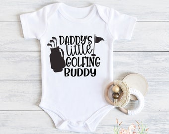 May include: White baby onesie with black text that reads "Daddy's little golfing buddy" and a black silhouette of a golf bag with clubs.