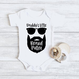May include: White baby onesie with black text and graphic. The text reads "Daddy's little beard puller". The graphic is a black silhouette of a man's face with sunglasses and a beard.